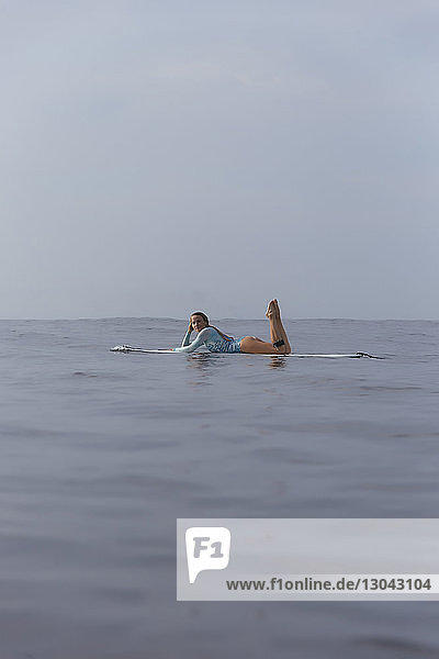 Seitenansicht einer sorgenfreien Frau  die auf einem Surfbrett im Meer gegen den Himmel liegt