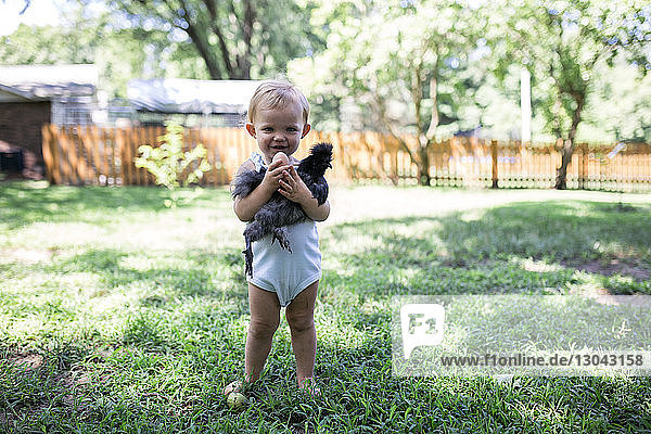 Portrait of cute baby girl holding chicken and egg while standing on grassy field at farm