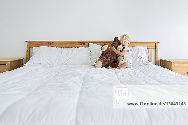 Porträt eines süßen Jungen  der einen Teddybär umarmt  während er auf dem Bett sitzt