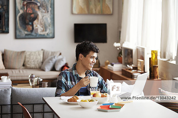 Glücklicher junger Mann benutzt Laptop beim Kaffeetrinken zu Hause