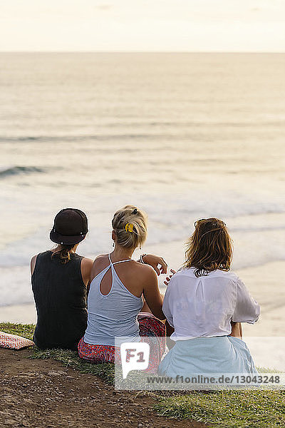 Rückansicht von Freundinnen  die bei Sonnenuntergang am Strand sitzen