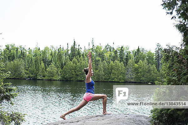 Seitenansicht einer Frau  die Yoga praktiziert  während sie auf einem Felsen am Seeufer vor klarem Himmel steht