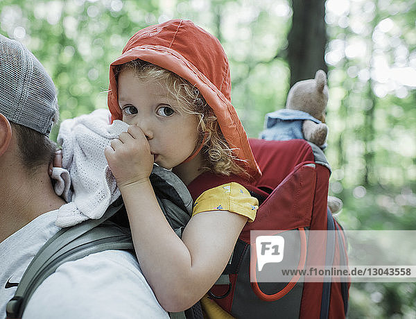 Portrait of cute daughter thumb sucking while being carried by father in forest