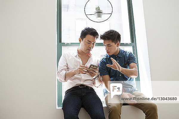 Kreative Geschäftsleute benutzen ein Smartphone  während sie im Büro auf dem Fensterbrett sitzen