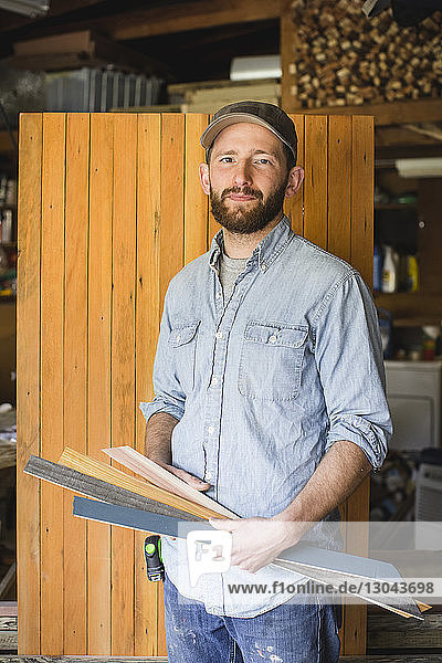 Portrait of man holding planks while standing against wood in workshop