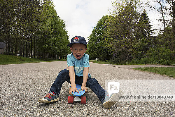Porträt eines verspielten Jungen  der auf einem Skateboard am Fussweg sitzt