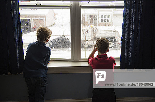 Rear view of brothers looking through window while standing at home