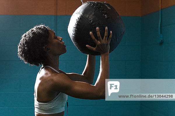 Side view of woman holding medicine ball while listening music against wall in gym