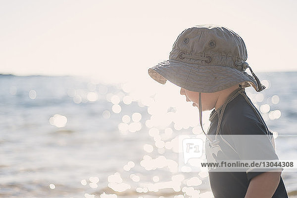 Seitenansicht eines Jungen mit Hut  der bei sonnigem Wetter am Strand gegen das Meer steht