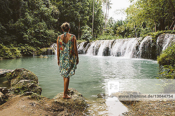 Rückansicht einer Frau  die auf einem Felsen vor Wasserfällen im Wald steht