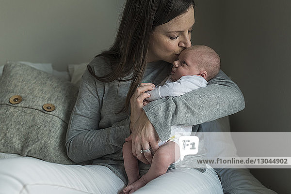 Mutter küsst neugeborenen Jungen  während sie zu Hause auf dem Sofa sitzt