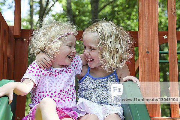 Happy sisters looking face to face while sitting on slide at playground