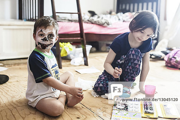 Porträt eines Jungen mit gruseliger Gesichtsbemalung,  der von seiner Schwester zu Hause auf dem Boden sitzt