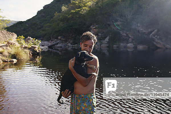 Shirtless man with dog standing in lake by mountain