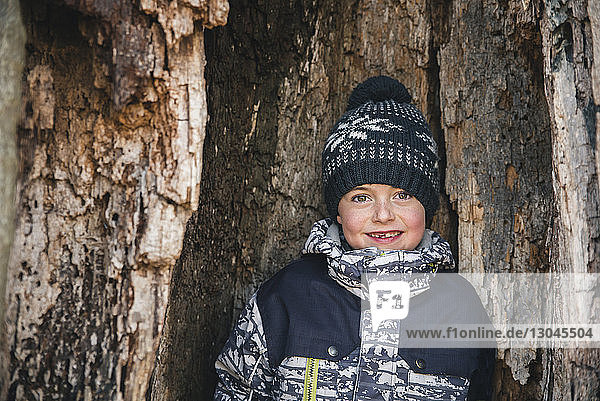 Porträt eines lächelnden Jungen  der im Winter im Wald am Baumstamm steht
