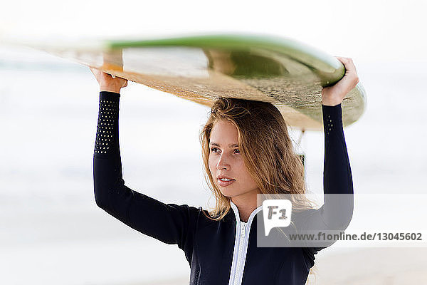 Frau schaut weg  während sie am Strand ein Surfbrett auf dem Kopf trägt