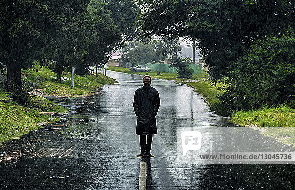 Junger Mann in voller Länge mit Händen in Regenmanteltaschen auf nasser Straße inmitten von Bäumen stehend