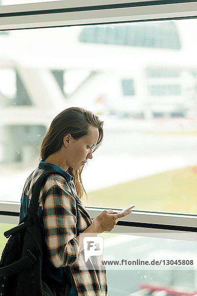 Frau benutzt Mobiltelefon  während sie am Fenster im Abflugbereich des Flughafens steht