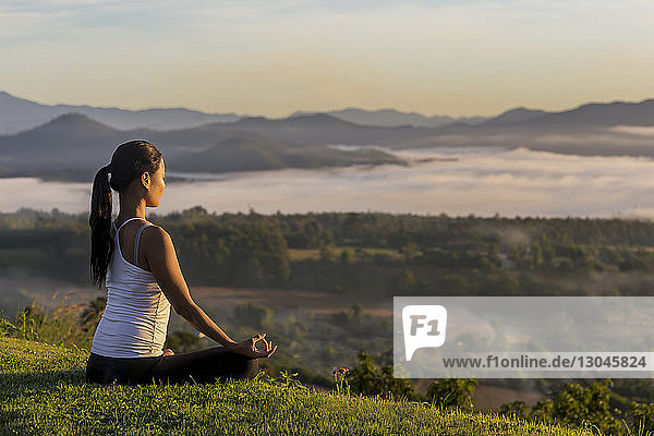 Seitenansicht einer Frau  die meditiert  während sie auf einem Grasfeld gegen den Himmel sitzt