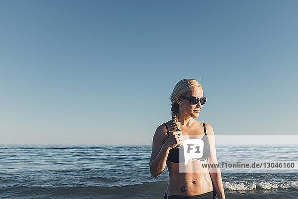 Nachdenkliche Frau mit Bikini und Sonnenbrille gegen Meer und klaren Himmel