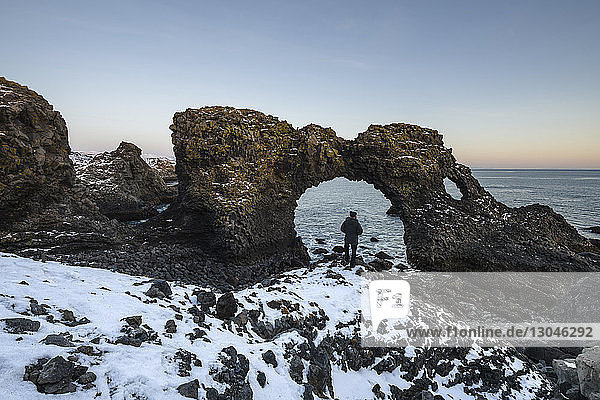 Rückansicht eines Wanderers der im Winter am natürlichen Bogen am Strand steht