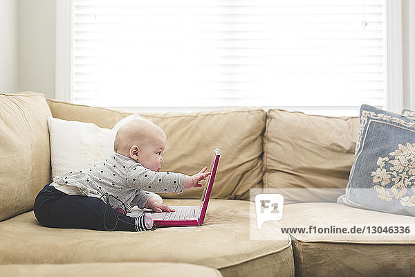 Side view of baby girl playing with laptop computer while sitting on sofa at home