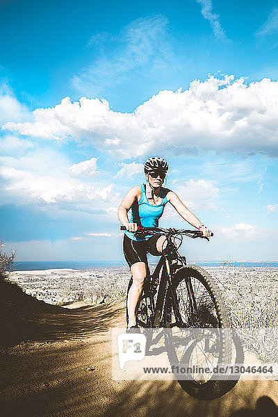 Full length of female cyclist riding bicycle on arid landscape against cloudy sky