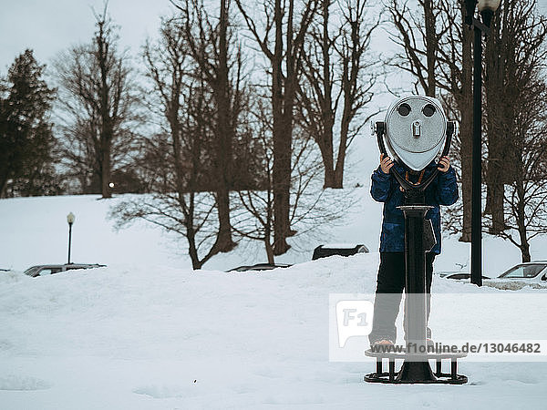 Boy looking through coin-operated binoculars on snowy field against bare trees
