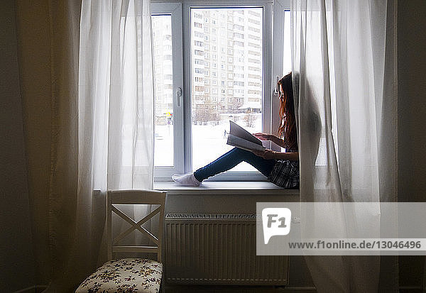 Side view of teenage girl reading book while sitting by window at home