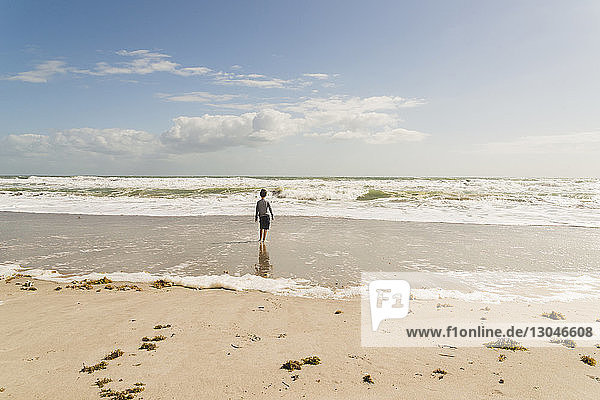 Rear view of boy standing at beach against sky during sunny day