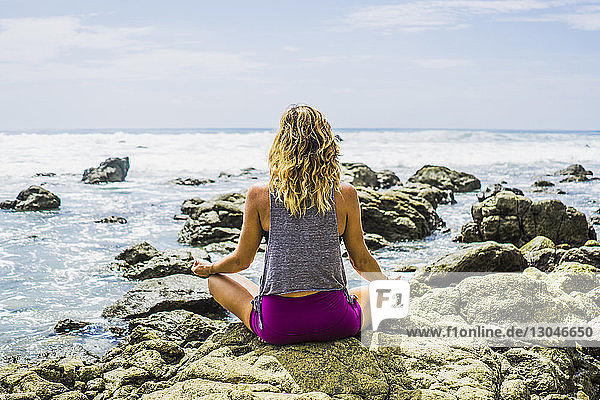 Rückansicht einer mittleren erwachsenen Frau  die am Strand auf Felsen meditiert