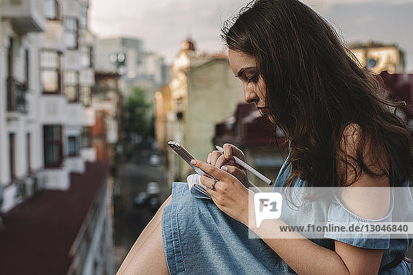Side view of young woman using smart phone while sitting against buildings in city
