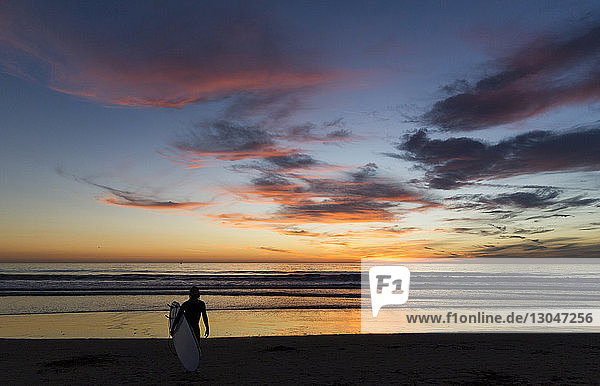 Rückansicht eines Mannes mit Surfbrett  der bei Sonnenuntergang am Strand gegen den Himmel läuft