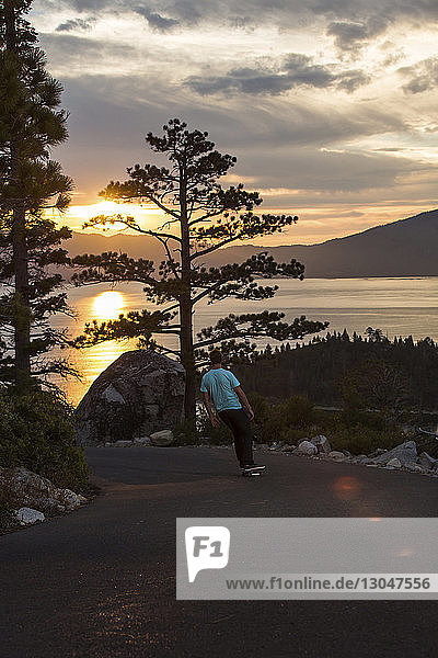 Mann fährt Skateboard auf Fußweg am See gegen den Himmel bei Sonnenuntergang