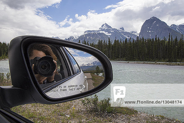 Reflection of man photographing with camera seen in side-view mirror of car against mountains