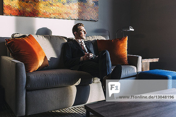 Nachdenklicher Geschäftsmann schaut weg  während er auf dem Sofa im Hotelzimmer sitzt
