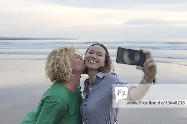 Fröhliche Frau  die sich mit einem Smartphone selbstständig macht  während sie bei Sonnenuntergang am Strand von einem Freund geküsst wird
