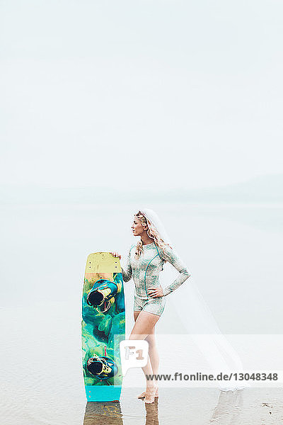 Frau trägt Schleier und hält Surfbrett  während sie bei nebligem Wetter am Strand gegen den Himmel steht