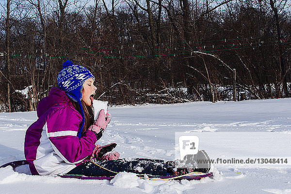 Side view of girl eating snow while sitting on field in forest