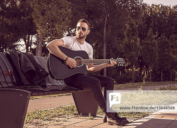 Ein Mann in voller Länge spielt Gitarre  während er an einem sonnigen Tag auf einer Parkbank sitzt