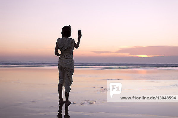 Rückansicht einer Frau  die mit einem Smartphone fotografiert  während sie bei Sonnenuntergang am Strand steht