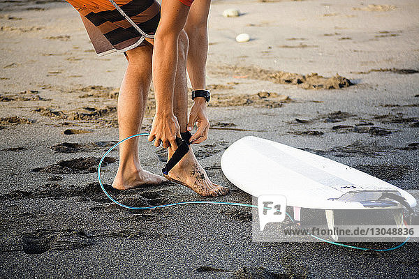 Low section of man tying surfboard while standing on wet sand at beach