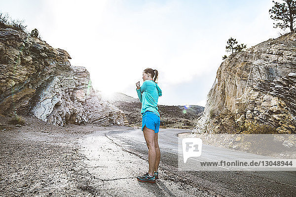 Seitenansicht einer Sportlerin in voller Länge  die die Arme ausstreckt  während sie auf einer Landstraße inmitten von Felsen steht