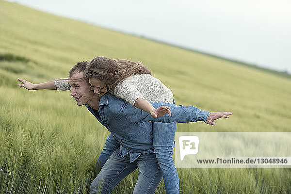 Fröhliches Paar mit ausgestreckten Armen genießt Huckepack-Ritt auf Grasfeld gegen den Himmel
