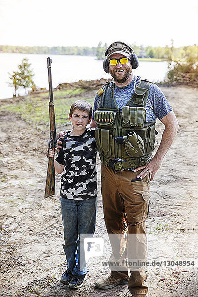 Portrait of son holding rifle while standing with father on field against sky