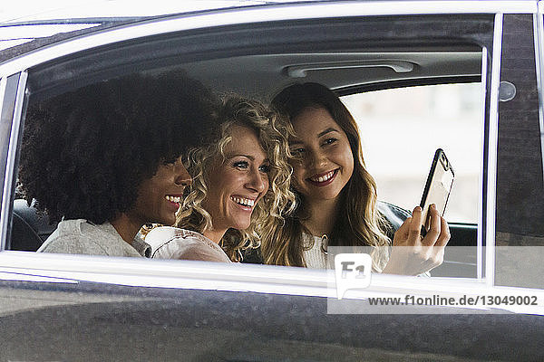 Fröhliche Geschäftsfrauen beim Selbermachen mit Smartphone im Auto durch Fenster gesehen