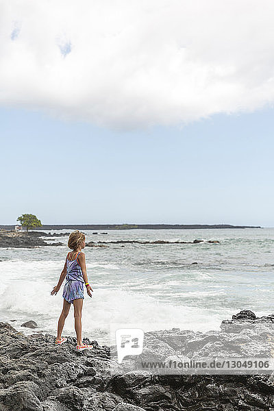 Rear view of girl standing on rocks at beach against cloudy sky