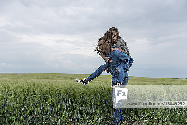Junger Mann nimmt Frau huckepack auf Grasfeld vor bewölktem Himmel