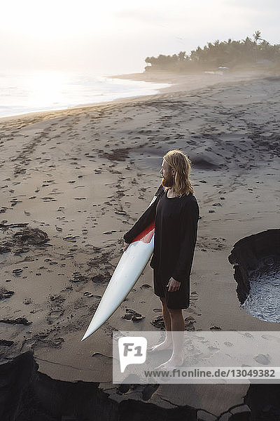 Mann in voller Länge schaut weg  während er ein Surfbrett am Strand trägt