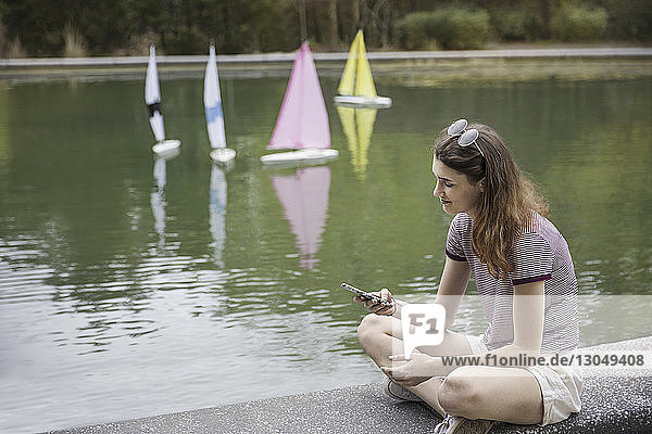 Teenager-Mädchen benutzt Smartphone  während sie auf einer Stützmauer am See im Park sitzt